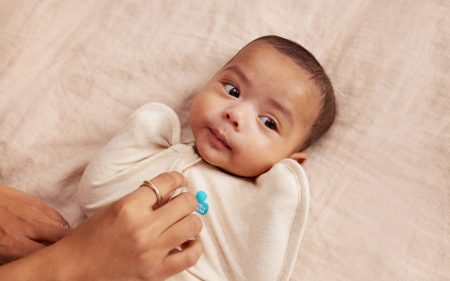 Baby lying on a soft surface in a beige swaddle up with a person's hand holdings settling them.